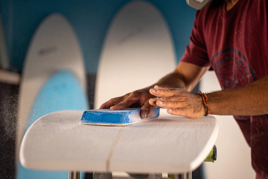 Shaper entrain de poncer une planche de surf dans son atelier. Gros plan sur main.
Crédit photo @dariotni